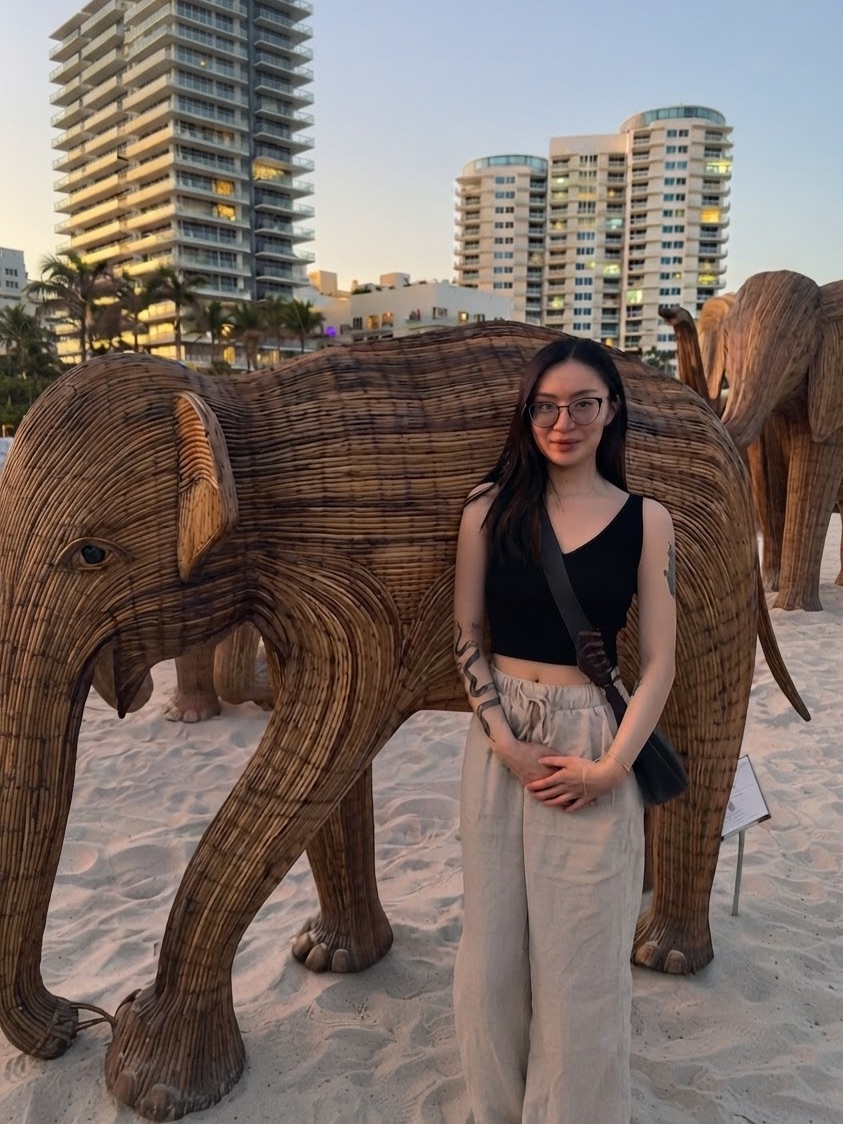 Yvonne Wu standing beside a woven elephant sculpture on a beach